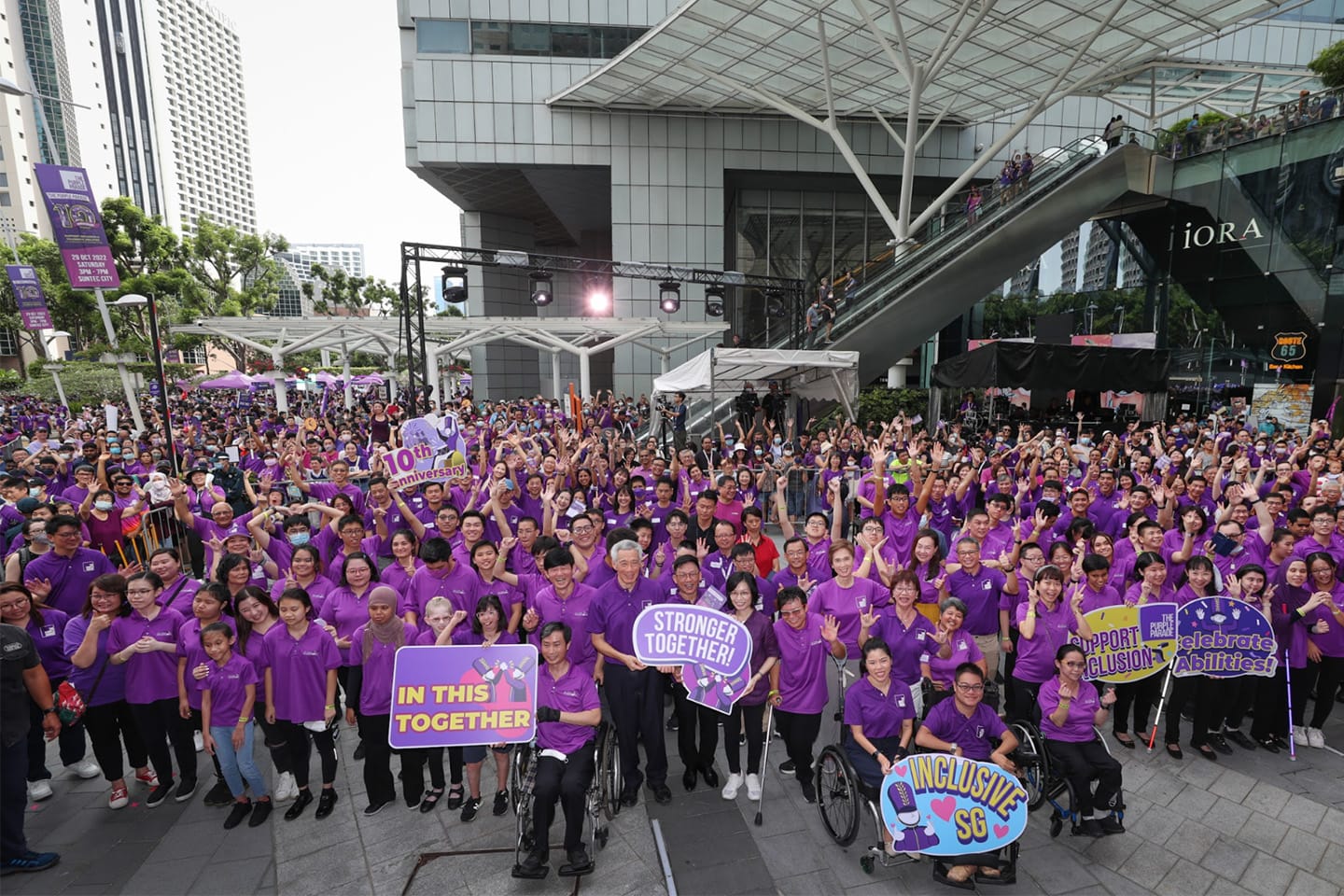 Large crowd in purple shirts holds signs. Two people in wheelchairs visible. Modern architecture backdrop.